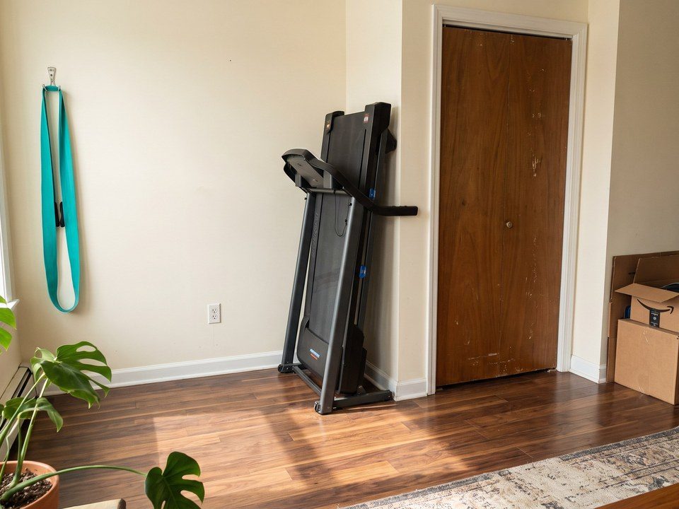 Folded treadmill standing against a wall by a door in a small apartment (illustrative AI image, not the exact product)